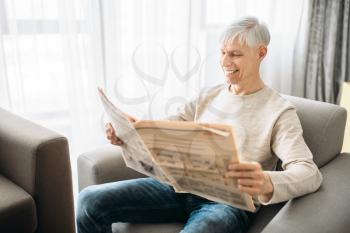 Adult man sitting on couch and reading newspaper at home. Mature male person relaxes in armchair