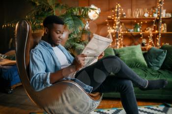 Black man reading newspaper in a comfortable leather chair in the living room, relaxation at home. Young african american male person in his house