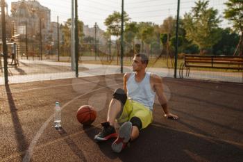 Basketball player sitting on the ground and drinks water on outdoor court. Male athlete in sportswear resting after streetball training
