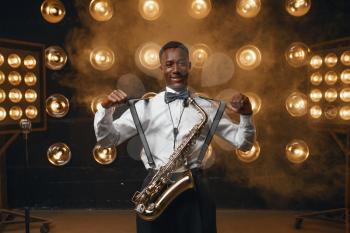 Black smiling jazz performer with saxophone on the stage with spotlights. Black jazzman preforming on the scene