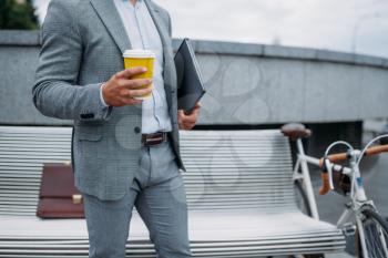 Businessman with cycle drinks coffee on the bench at the office building in downtown. Business person riding on eco transport on city street, urban style