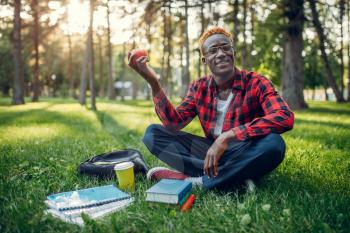 Black student with apple sitting on the grass in summer park. A teenager studying outdoors and having lunch