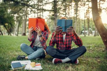Students sitting on the grass and cover their faces with books, summer park. Male and female teenagers studying outdoors and having lunch