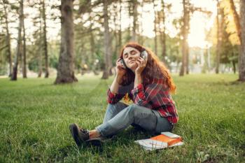Female student in headphones sitting on the grass in summer park. Redheaded teenager studying and leisures outdoors