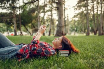 Female student with phone lying on the grass in summer park. Redheaded teenager studying and leisures outdoors
