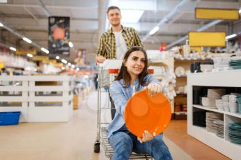 Young couple jokes with cart and plate in houseware store. Man and woman buying home goods in market, family in kitchenware supply shop
