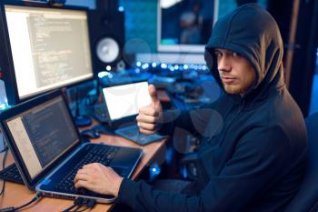 Hacker in hood shows thumbs up at his workplace with laptop and PC, password or account hacking. Internet spy, crime lifestyle, risk job, network criminal