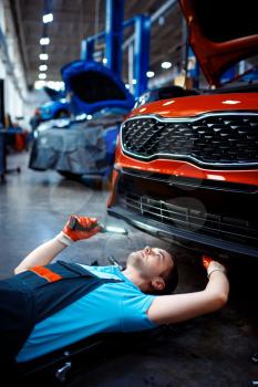 Worker in uniform lying under the vehicle, car service station. Automobile checking and inspection, professional diagnostics and repair