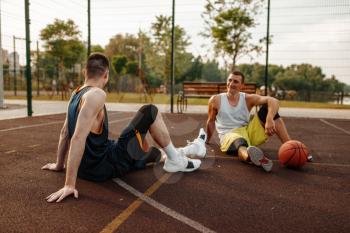 Two basketball players sitting on the ground on outdoor court. Male athletes in sportswear after streetball training