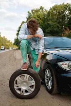 Car breakdown, young man puts the spare tyre. Broken automobile or problem with vehicle, trouble with punctured auto tire on highway
