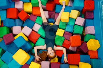 Cute little girl lying in soft cubes, playground in entertainment center. Play area indoors, playroom