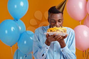 Cheerful man in cap tasting birthday cake, yellow background. Smiling male person got a surprise, event celebration, balloons decoration