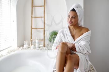 Attractive woman in bathrobe sitting near the bath. Female person in bathtub, beauty and health care in spa, wellness treathment in bathroom, pebbles and candles on background