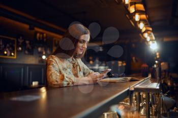 Pretty woman with mobile phone sitting at the counter in bar. One female person in pub, human emotions, leisure activities, nightlife
