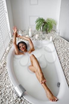 Young happy woman, relaxation in bath with milk. Female person in bathtub, beauty and health care in spa, wellness treathment in bathroom, pebbles and candles on background