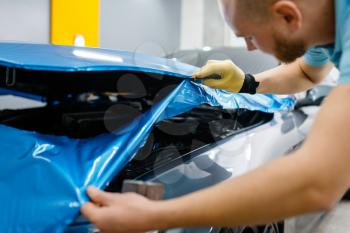 Car wrapping, man cuts protective vinyl foil or film on vehicle hood closeup. Worker makes auto detailing. Automobile paint protection coating, professional tuning