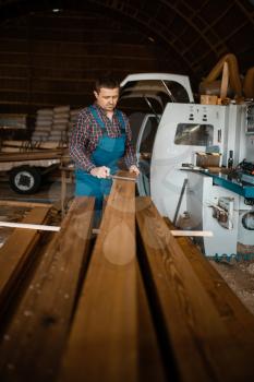 Carpenter in uniform measures the board with caliper, woodworking machine on background, lumber industry, carpentry. Wood processing on factory, forest sawing in lumberyard