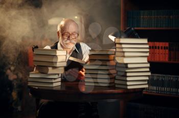 Elderly writer sitting at the table with stack of books in home office. Old man in glasses writes literature novel in room with smoke, inspiration