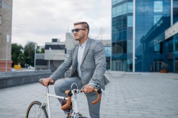 One businessman in sunglasses poses on bicycle at the office building in downtown. Business person riding on eco transport on city street