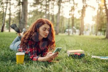 Female student with cup of coffee lying on the grass in summer park. Ginger teenager studying and leisures outdoors