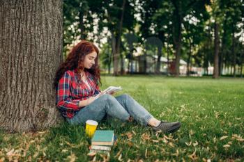 Female student studying the book on the grass in summer park. Ginger teenager leisures outdoors, pretty girl relax outside