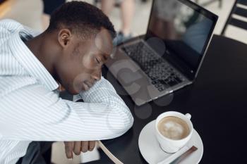 Young ebony businessman with laptop sleeping in office cafe. Tired business person drinks coffee in food-court, black man in formal wear