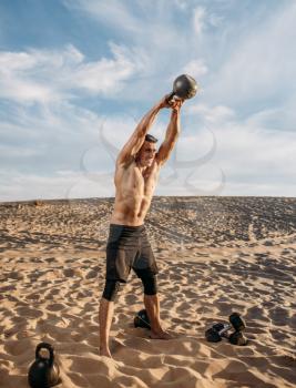 Muscular male athlete doing exercises with kettlebell in desert at sunny day. Strong motivation in sport, strength outdoor training, sportsman with weights