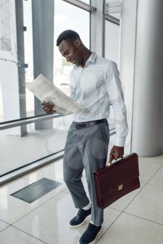 Black businessman with briefcase reading newspaper, mall. Successful business person, black man in formal wear, shopping center
