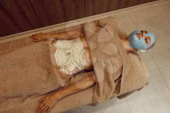 Slim woman with cream mask on her face lying on massage table, top view. Massaging and relaxation, body and skin care. Female person relaxing in spa salon