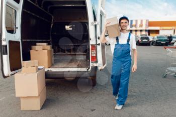 Deliveryman in uniform poses at the car with parcel boxes, delivery service. Man standing at cardboard packages in vehicle, male deliver, courier or shipping job