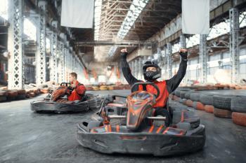 Go kart racer in helmet raised his hands up, winner, front view, karting auto sport indoor. Speed race on close go-kart track with tyre barrier. Fast vehicle competition, hot pursuit