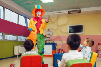 Funny clown juggles in front of surprised children. Birthday party celebrating in playroom, baby holiday in playground. Childhood happiness, childish leisure
