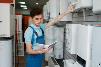 Plumber in uniform choosing boiler at showcase in plumbering store. Man with notebook buying sanitary engineering in shop, water heater choice