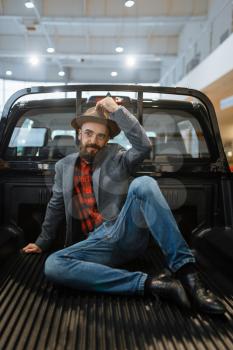 Smiling man poses in the back of new pickup truck in car dealership. Customer in vehicle showroom, male person buying transport, auto dealer business