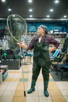 Male angler in rubber jumpsuit holds net in fishing shop, hooks and baubles on background. Fisherman buying equipment and tools for fish catching and hunting, assortment on showcase in store