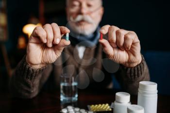 Elderly man holds pills in his hands, home office on background, age-related diseases. Mature senior is ill and being treated in his house