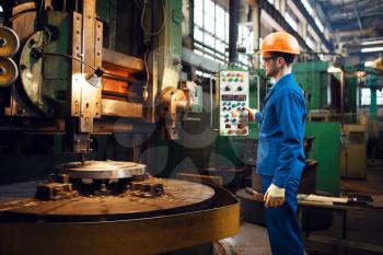 Turner in uniform and helmet works on large lathe, factory. Industrial production, metalwork engineering, power machines manufacturing