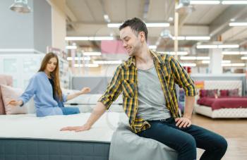 Young love couple checks mattress in furniture store showroom. Man and woman looking samples for bedroom in shop, husband and wife buys goods for modern home interior