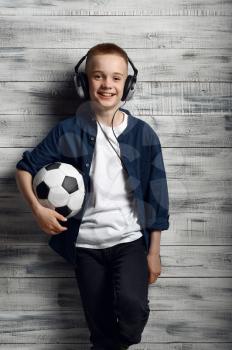 Little boy in headphones holds ball in studio. Children and gadgets, kid isolated on wooden background, child emotion, schoolboy photo session