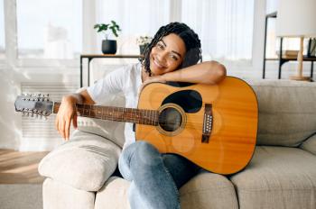 Happy woman with guitar sitting on sofa at home. Pretty lady in earphones relax in the room, female sound lover resting