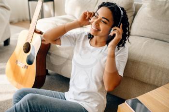 Smiling woman in headphones at home, guitar on background. Pretty lady with musical instrument relax in the room, female music lover resting