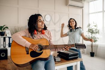 Funny women play the guitar and listen to music at home. Pretty girlfriends in earphones relax in the room, sound lovers resting on couch, female friends leisures together