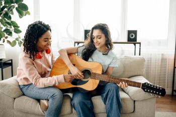 Women learn to play the guitar at home. Pretty girlfriends in earphones relax in the room, sound lovers resting on couch, female friends leisures together