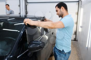 Male worker tries on wetted car tinting, tuning service. Mechanic applying vinyl tint on vehicle window in garage, tinted automobile glass