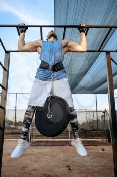 Muscular man does pull-ups exercise with weight on a horizontal bar, street workout, crossfit. Fitness training on sports ground outdoor, male person pumps muscles, active urban lifestyle