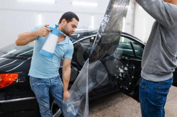 Male worker with spray wetting car tinting, tuning service. Mechanic applying vinyl tint on vehicle window in garage, tinted automobile glass