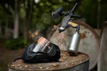 Paintball gun and protection mask closeup, nobody, playground in the forest on background. Extreme sport outdoors, pneumatic weapon and paint bullets, military team game concept