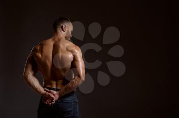 Strong male athlete poses in studio, back view, dark background. One man with athletic build, shirtless sportsman in jeans pants, active healthy lifestyle