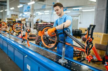 Bicycle factory, worker at assembly line, wheel installation. Male mechanic in uniform installs cycle parts in workshop, industrial manufacturing