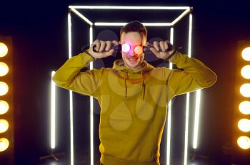 Young gamer holds virtual reality gamepads at his eyes in luminous cube, front view. Dark playing club interior, spotlight on background, VR technology with 3D vision